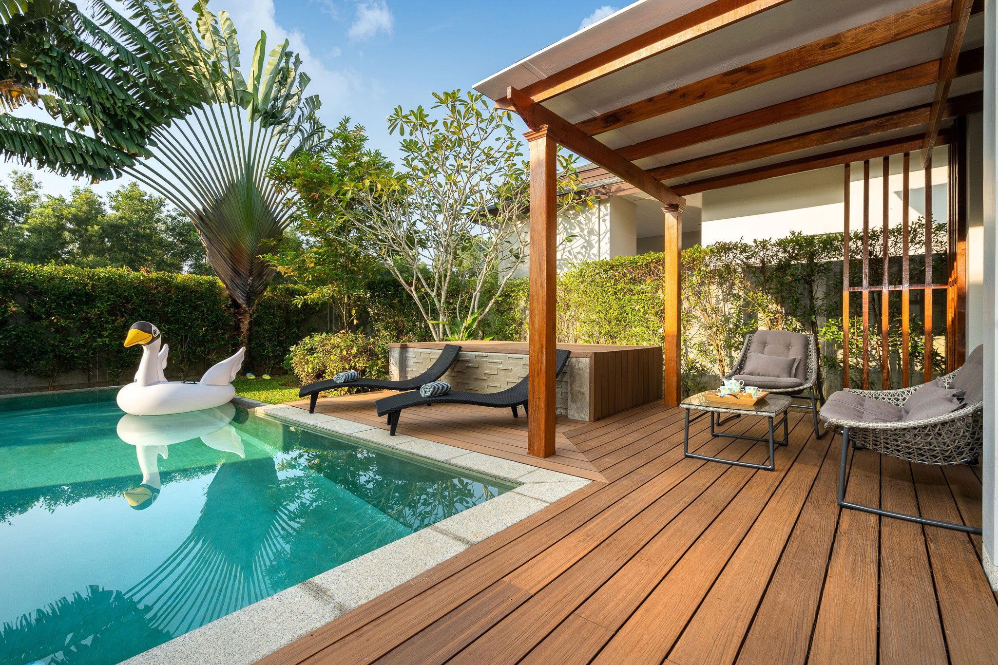 A backyard with a swimming pool featuring a swan-shaped float, wooden deck with lounge chairs, small table, two chairs, a banana tree, and lush greenery surrounding the area.